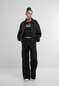 Jachetă bomber din suede negru peste un tricou croppat negru cu logo, asortată cu pantaloni cargo negri, largi, și adidași albi.