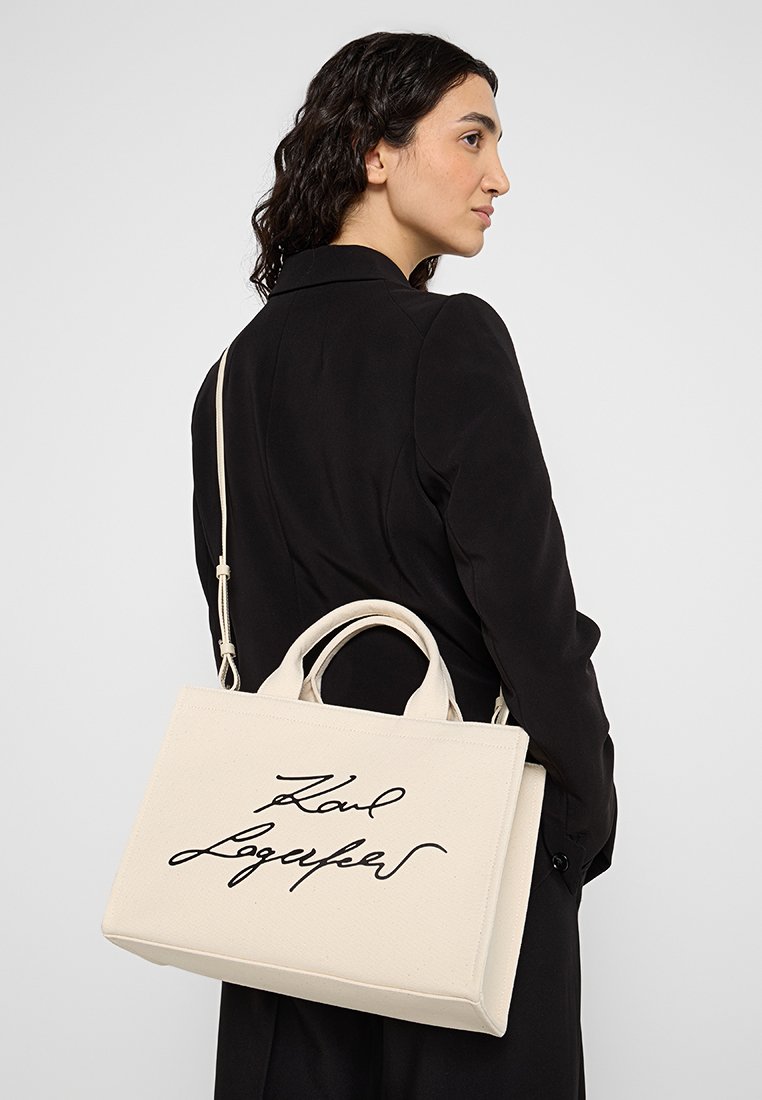 Femme en manteau noir portant un sac fourre-tout beige Karl Lagerfeld avec une bandoulière sur le dos, contre un fond gris.