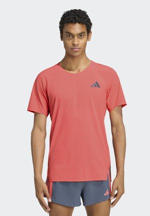 Młody mężczyzna o kręconych włosach, ubrany w koralowy sportowy t-shirt i szare szorty z koralowymi paskami, stojący na jednolitym jasnym tle.