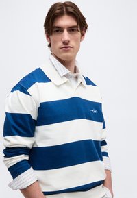 Νεαρός άνδρας με καστανά μαλλιά μέσου μήκους, φορώντας μπλούζα ράγκμπι με μπλε και λευκές ρίγες πάνω από λευκό πουκάμισο με γιακά.