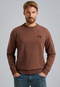 Sweatshirt marron à manches longues et col rond, avec une petite étiquette sur la poitrine. Fabriqué en tissu doux, avec des poignets et un ourlet côtelés.