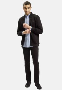 Czarna kurtka zapinana na zamek nad jasnoniebieską koszulą, zestawiona z czarnymi dżinsami i ciemnymi tenisówkami. Gładka tekstura materiału z prążkowanymi mankietami.