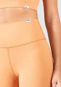 Soutien-gorge de sport de couleur pêche et leggings taille haute en tissu lisse et extensible avec des étiquettes de marque contrastantes près de la taille.