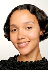 Portrait d'une personne avec des cheveux ondulés, de longueur moyenne et foncés, portant un blouse noire texturée. Peau lisse et un sourire subtil.