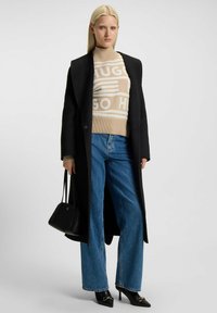 Zwarte jas over een beige trui met logo, blauwe wijde jeans en zwarte hakken. Kleine zwarte handtas en lang steil haar.