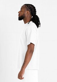 Vit kortärmad t-shirt med avslappnad passform. Mjuk tyg, rund halsringning och rak fåll. Sido vy som visar flätad frisyr och tatueringar.