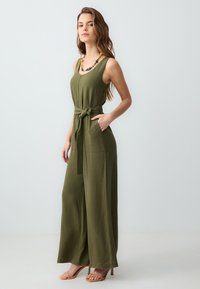 Femme portant une combinaison sans manches vert olive, avec des jambes larges, une ceinture nouée à la taille et un collier en perles colorées, se tenant devant un fond blanc.