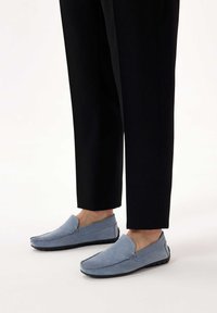 Light blue suede slip-on shoes with a flat black sole and stitch detailing, paired with black tailored pants, showing a casual look.