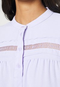 Blusa lilla chiara con colletto alla mandarino, chiusura a bottoni e dettagli in pizzo decorativo sul petto. Tessuto morbido con plissé.