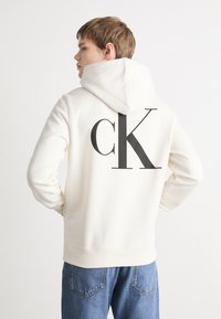 Sudadera crema con un gran logo "ck" en gris oscuro en la espalda, con capucha ajustable y puños acanalados, combinada con vaqueros azules.