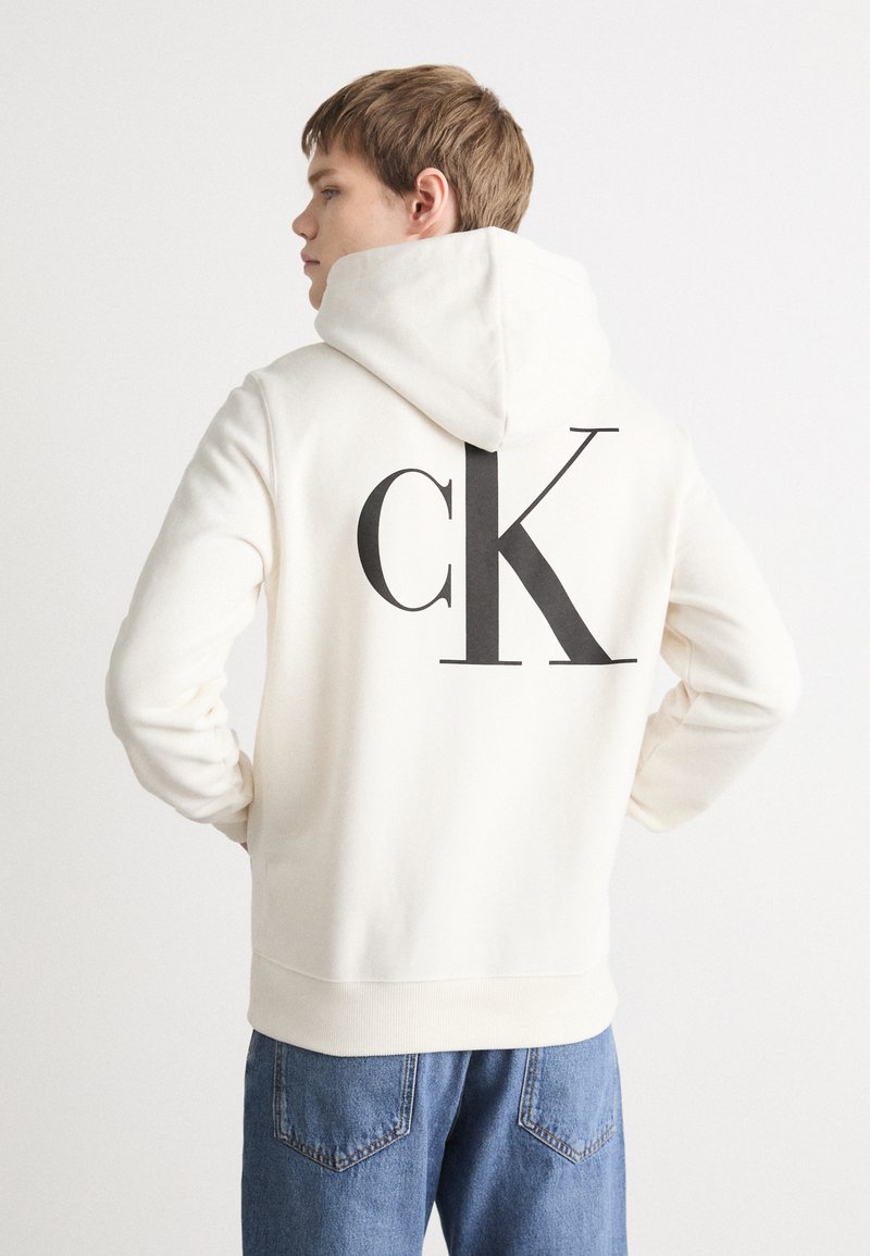 Sudadera crema con un gran logo "ck" en gris oscuro en la espalda, con capucha ajustable y puños acanalados, combinada con vaqueros azules.