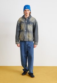 Denimjakke med blege blå nuancer, klassisk design, knaplukning, to forlommer. Båret med løstsiddende blå jeans og en blå hue.