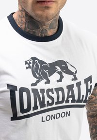 Valkoinen puuvillateepaita, jossa musta leijonagrafiikka ja "LONSDALE LONDON" -teksti. Pyöreä sininen kaulus ja lyhyet hihat, näkyvissä tekstuuri-tatuointi.
