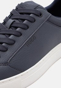 Σκούρο μπλε δερμάτινο αθλητικό παπούτσι με κορδόνια στο ίδιο χρώμα και λευκή λαστιχένια σόλα, με ανάγλυφο λογότυπο "HUGO" στην πλαϊνή επιφάνεια.