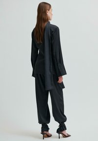 Černá halenka s dlouhými rukávy a zadním drapem, v kombinaci s volnými černými kalhotami. Texturovaná látka a černé sandály na vysokém podpatku se řemínky.