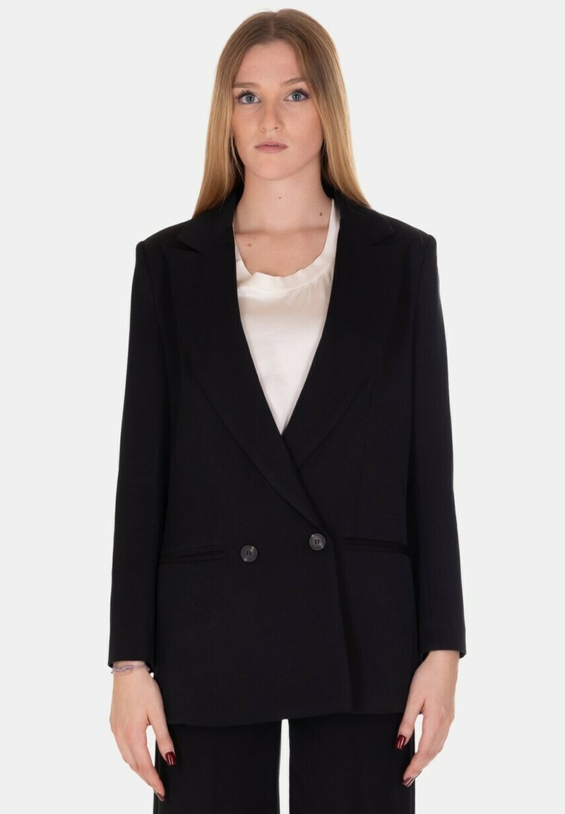 Blazer doppiopetto nero con una vestibilità sartoriale, rever a punta, due bottoni frontali e un tessuto texturizzato. Indossato sopra una blusa bianca.