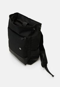 Svart stoff tote bag med en glidelåslomme foran, nett skulderrem og en glatt bunnpanel. Har trykknapplukking og et enkelt design.