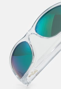 Occhiali da sole con montatura trasparente, lenti verde sfumate verso il blu e un discreto logo dorato sul lato.