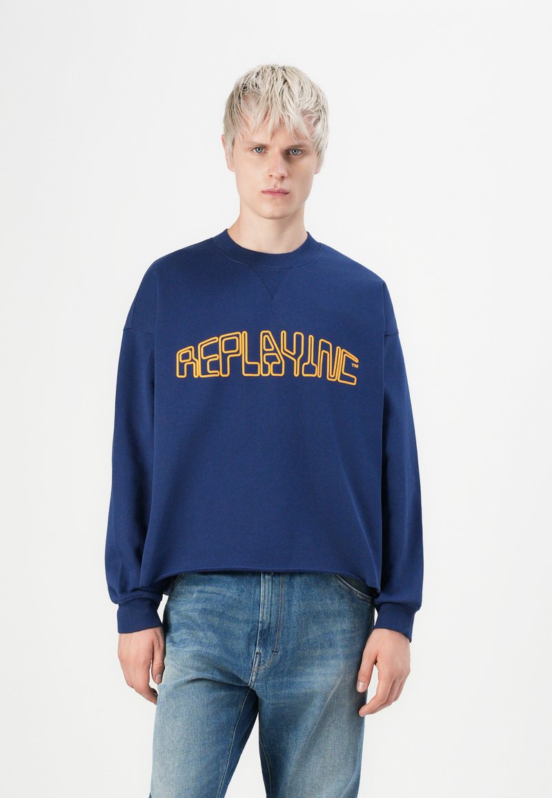 En blond person som har på seg en løs, marineblå sweatshirt med gul tekst "REPLAYING" og lyseblå jeans, står mot en hvit bakgrunn.