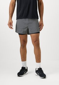 Pantalones cortos deportivos grises con un ribete negro, presentan una cinturilla elástica, bolsillos laterales y un pequeño logo. Se llevan con una camiseta oscura y zapatillas.