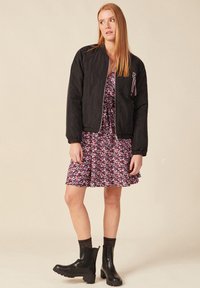 Zwarte bomberjack met een gladde textuur, voorzien van geribde manchetten en een kleurrijke accentlint op de borst, gecombineerd met een bloemenjurk en zwarte laarzen.