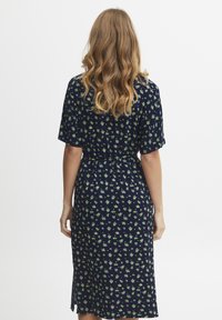 Marinblå klänning med korta ärmar, blomm�önster i grönt, elastisk midja och sidoflikar för lätt rörelse. Mjukt tyg med lätt textur.