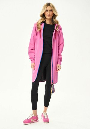 Kobieta stojąca w długiej różowej kurtce z niebieskim zamkiem, czarnych legginsach, czarnej bluzce i różowych trampkach na białym tle.