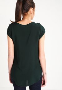 Žena s tmavohnědým culíkem, oblečená v tmavě zeleném tričku s krátkým rukávem a tmavých kalhotách, viděná zezadu na bílém pozadí.