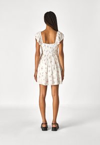 Vit blommig klänning med korta volangärmar, fyrkantig rygg och elastisk midja. Fläktande kjol med samlade lager, bärs med svarta skor.