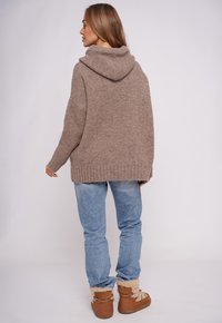 Beżowa bluza z kapturem o teksturowanym splocie, luźnym kroju i ściągaczu na dole, zestawiona z jasnoniebieskimi dżinsami oraz brązowymi butami ocieplanymi futerkiem.