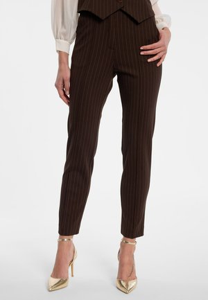 Vrouw draagt donkerbruine, op maat gemaakte pantalon met krijtstreep, een bijpassend gilet en gouden hoge hakken met enkelbandje, staand met één hand op de heup.