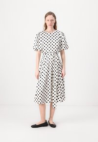 Vestido branco com bolinhas pretas, mangas curtas em balão, cinto na cintura, saia fluida. Usado com sapatos flats pretos, em um fundo simples.