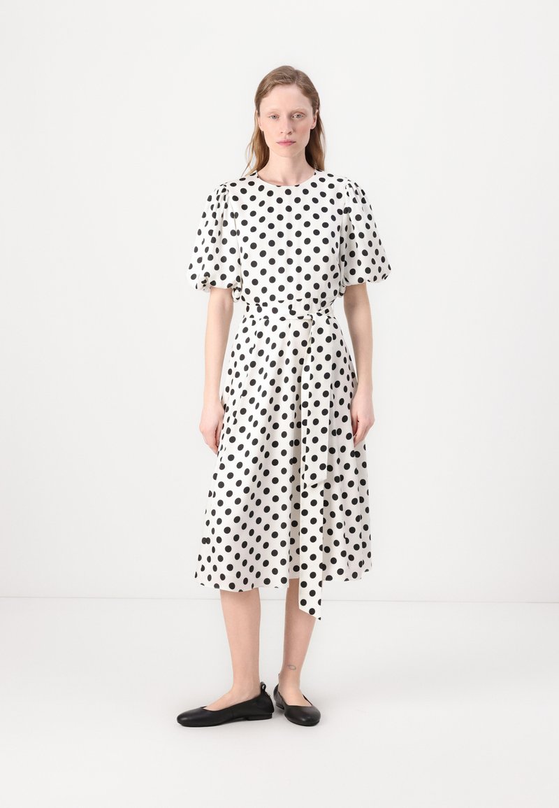 Vestido branco com bolinhas pretas, mangas curtas em balão, cinto na cintura, saia fluida. Usado com sapatos flats pretos, em um fundo simples.