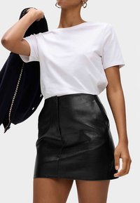 T-shirt blanc à manches courtes associé à une mini-jupe en cuir noire avec une fermeture éclair à l'avant et une texture lisse et brillante. Une veste bleu marine est posée sur le bras.