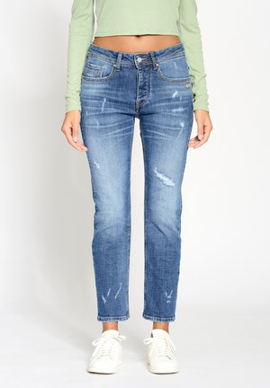 Personne portant un crop top à manches longues vert clair, un jean bleu déchiré et des baskets blanches, debout sur un fond blanc uni.