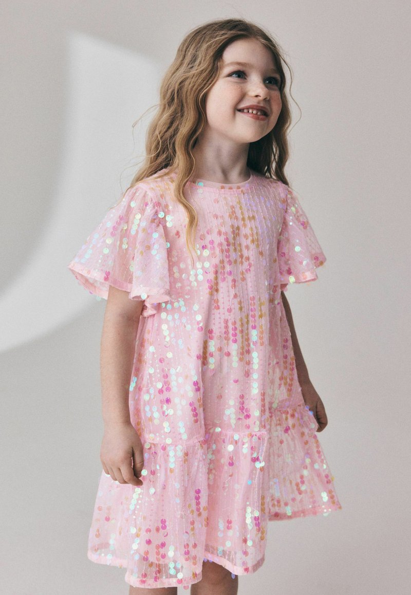 Jeune fille souriante aux longs cheveux ondulés portant une robe rose couverte de sequins irisés, debout devant un fond uni.