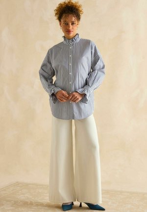 Svītrains krekliņš ar viļņotu apkakli, kombinēts ar platām krēma krāsas biksēm un zilām norādītām kurpēm ar augstiem papēžiem. Gluda auduma, brīva piegriezuma.