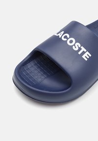 Chausson en plastique bleu marine avec un design à bout ouvert. Présente une semelle intérieure texturée avec un motif en grille et un logo blanc sur la lanière.