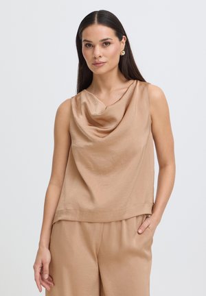 Femme aux longs cheveux foncés portant un haut sans manches en satin beige avec col bénitier et un pantalon assorti, debout avec une main dans la poche.