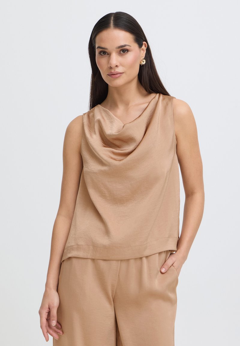 Femme aux longs cheveux foncés portant un haut sans manches en satin beige avec col bénitier et un pantalon assorti, debout avec une main dans la poche.