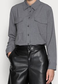 Grå långärmad skjorta med knäppning och krage samt två bröstfickor, matchad med höga svarta byxor i konstläder. Slät textur.