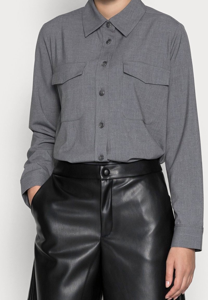 Grå långärmad skjorta med knäppning och krage samt två bröstfickor, matchad med höga svarta byxor i konstläder. Slät textur.