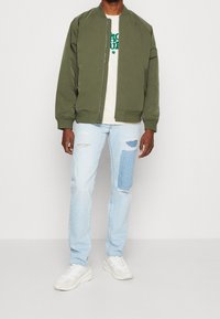 Grön bomberjacka, beige skjorta med grön text, ljusblå slitna jeans med blå patch och vita sneakers. Enkel och avslappnad design.