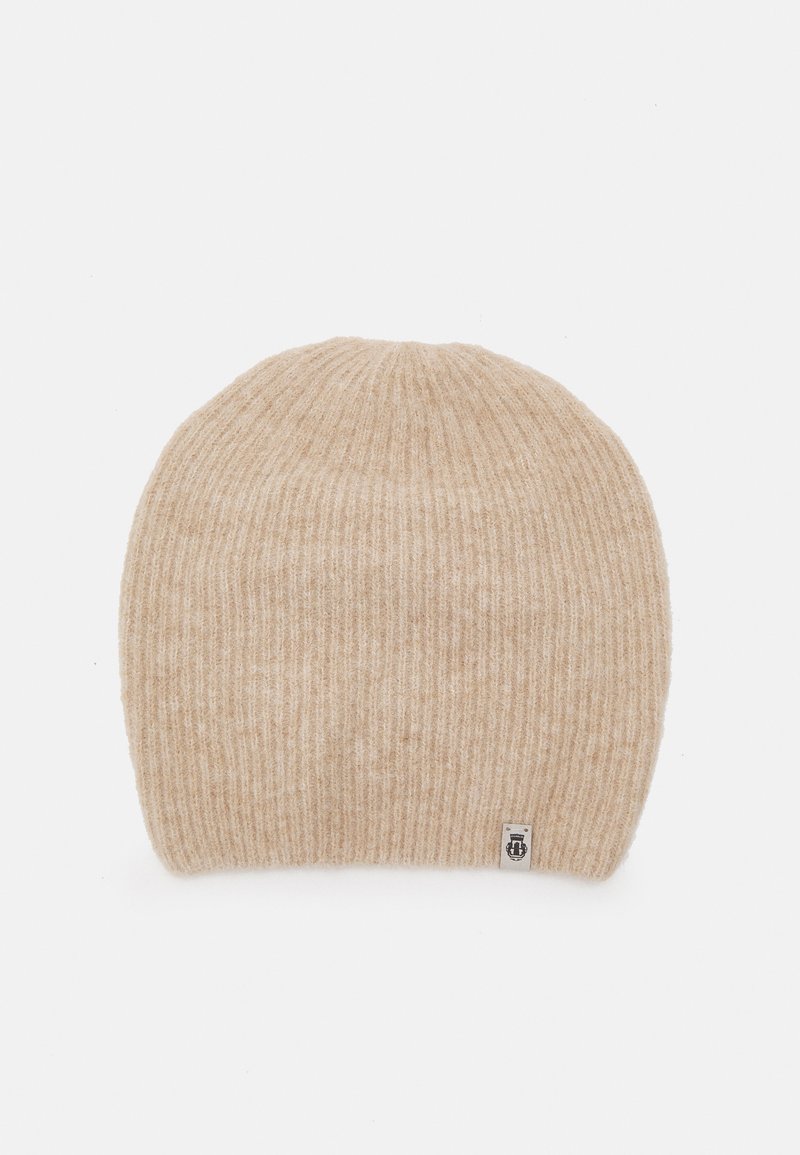 Beige knitted beanie with a ribbed texture, featuring a small logo tag on the side. Soft material, rounded top design.