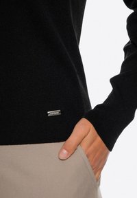Czarny sweter dziany o prążkowanej fakturze, z małą srebrną metką logo na dolnym brzegu. Ręka oparta w beżowej kieszeni.