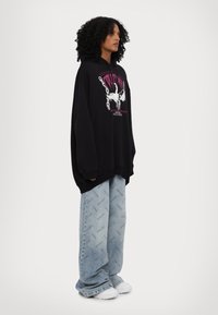 Black oversized hoodie with graphic design, paired with light blue jeans featuring repeated text pattern. White sneakers complete the outfit.