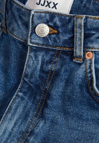 Jean en denim avec un lavage bleu foncé, des coutures claires, un bouton en argent et une étiquette marquée "JJXX" sur la taille.