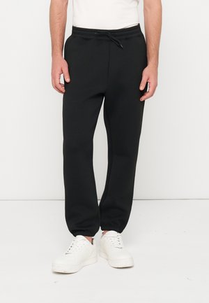 Mann, der schwarze Jogginghose mit Kordelzug und weiße Sneakers trägt, steht vor einem einfachen weißen Hintergrund.