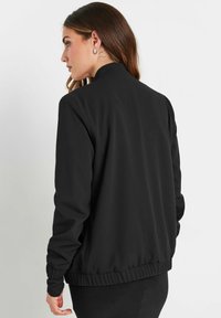 Czarna kurtka bombowiec o gładkiej teksturze, z prążkowanymi mankietami i dołem oraz stójką. Prosty design bez widocznych logo i wzorów.