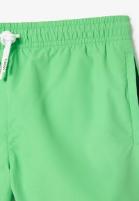 Lichtgroene zwembroek met elastische tailleband, witte koord in een knoop gestrikt, en een zijzak zichtbaar aan de rechterkant.